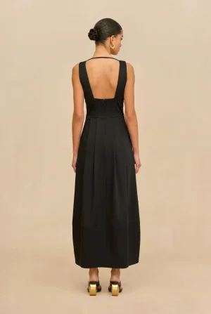 Back view of a woman in a black sleeveless gown with an open back, braided bun, and gold earrings, wearing gold-heeled sandals.