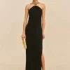 Model wearing a long black halter-neck gown with a gold neckpiece and matching gold clutch, standing against a beige background.