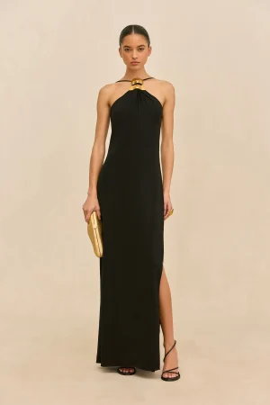 Model wearing a long black halter-neck gown with a gold neckpiece and matching gold clutch, standing against a beige background.