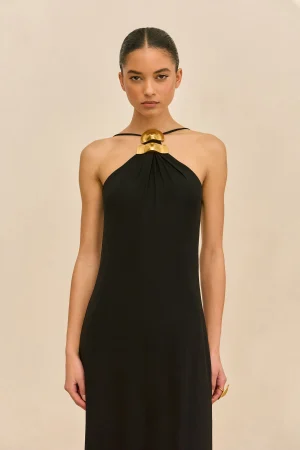 Model wearing a black halter dress with a bold gold geometric necklace, standing against a beige background.