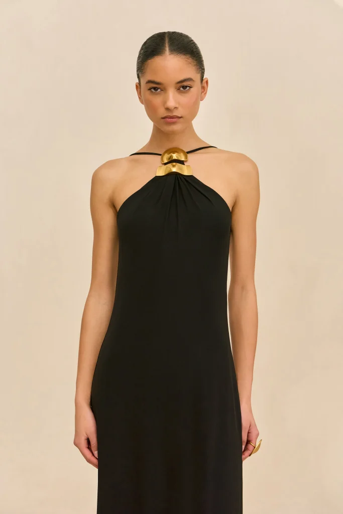 Model wearing a black halter dress with a bold gold geometric necklace, standing against a beige background.
