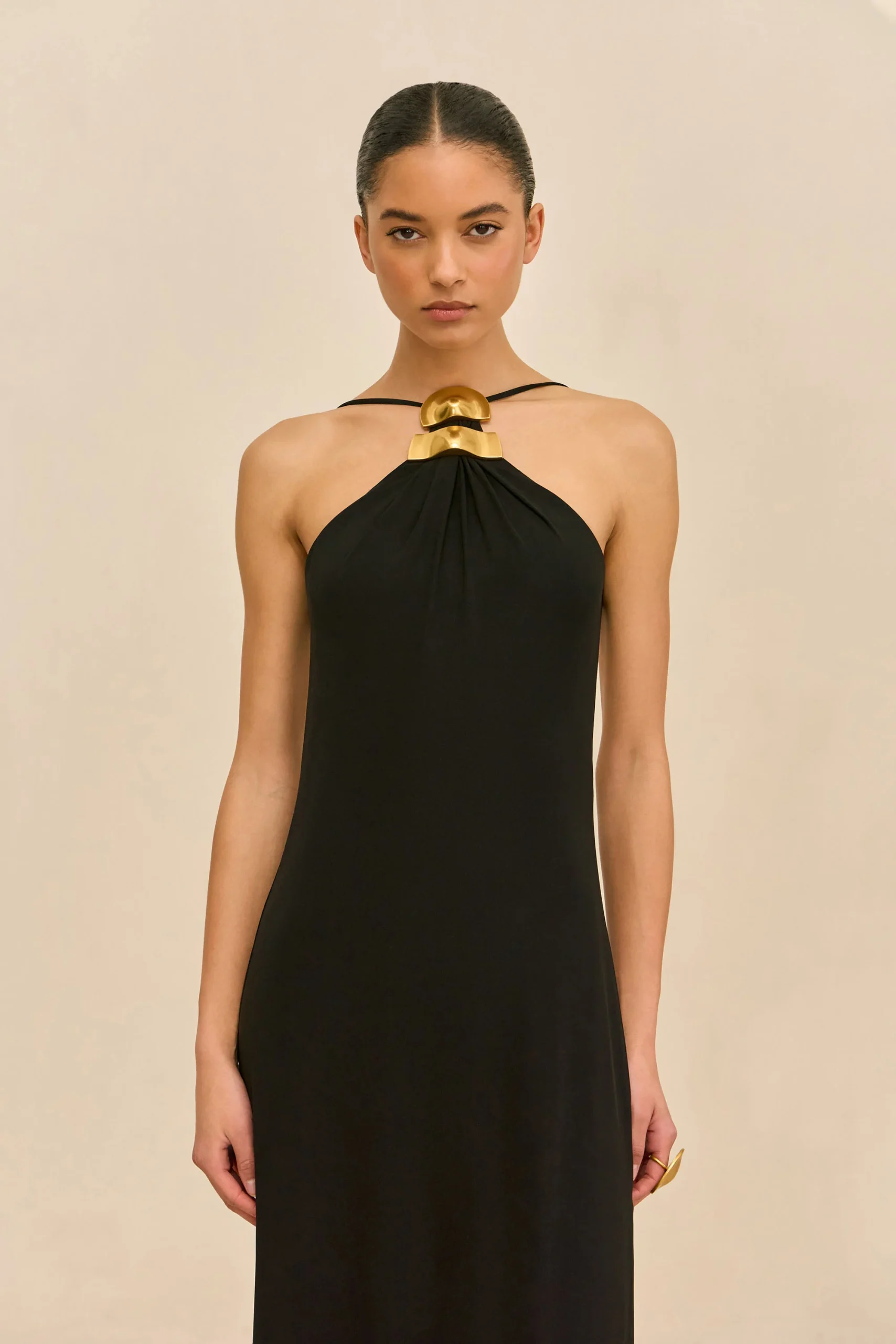 Model wearing a black halter dress with a bold gold geometric necklace, standing against a beige background.
