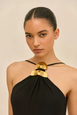 Portrait of a woman with slicked-back hair wearing a black halter dress and large gold statement necklace against a beige background, looking at the camera.