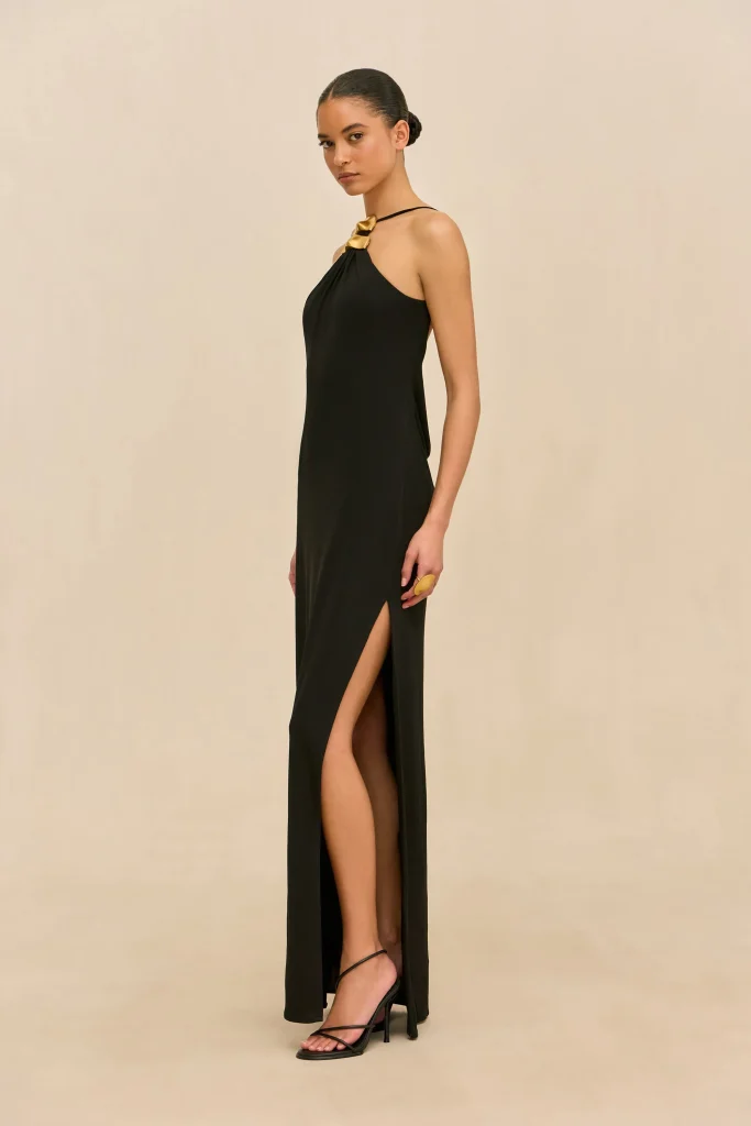 Full-length image of a woman in a black halter gown with a high slit, gold bow necklace, and black strappy heels against a beige backdrop, posed in profile.
