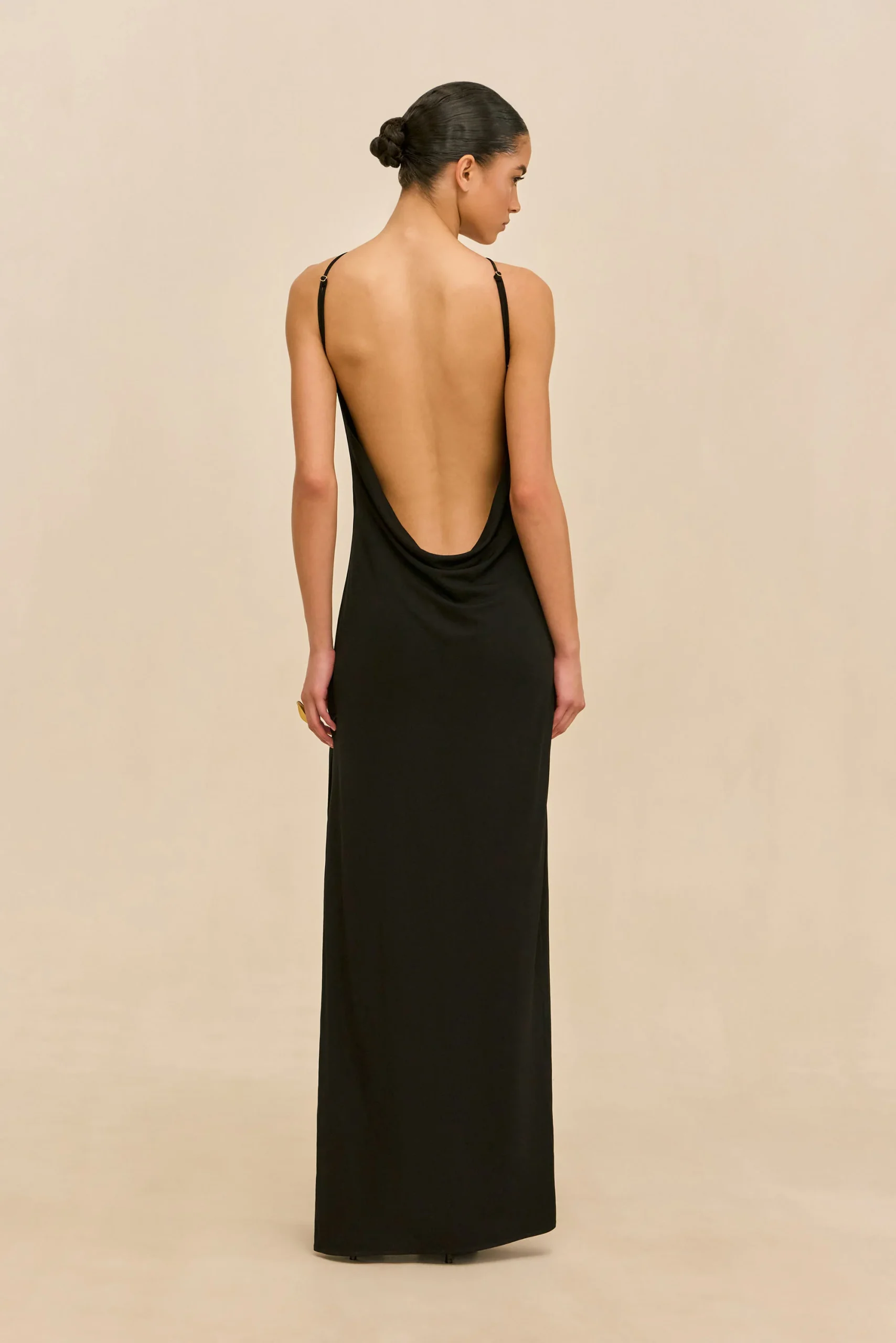 Woman in a long black backless dress, viewed from behind, with a neat bun in her hair against a beige backdrop.