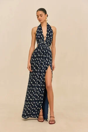 Woman wearing a navy halter maxi dress with white polka dots, cutouts at the waist, and a high slit, paired with gold earrings and brown sandals.