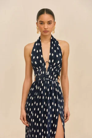 Female model wearing a navy halter-neck dress with white polka dots, standing against a pale beige background, with gold sculptural earrings and a high-sleek hairstyle.