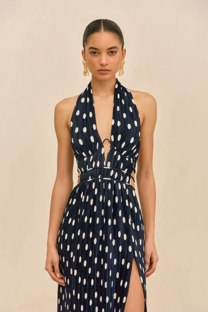 Female model wearing a navy halter-neck dress with white polka dots, standing against a pale beige background, with gold sculptural earrings and a high-sleek hairstyle.