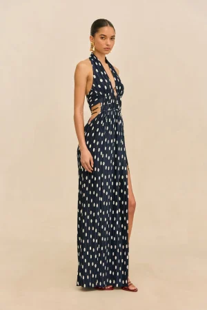Model wearing a navy polka-dot halter maxi dress with side cutouts, standing against a beige backdrop.