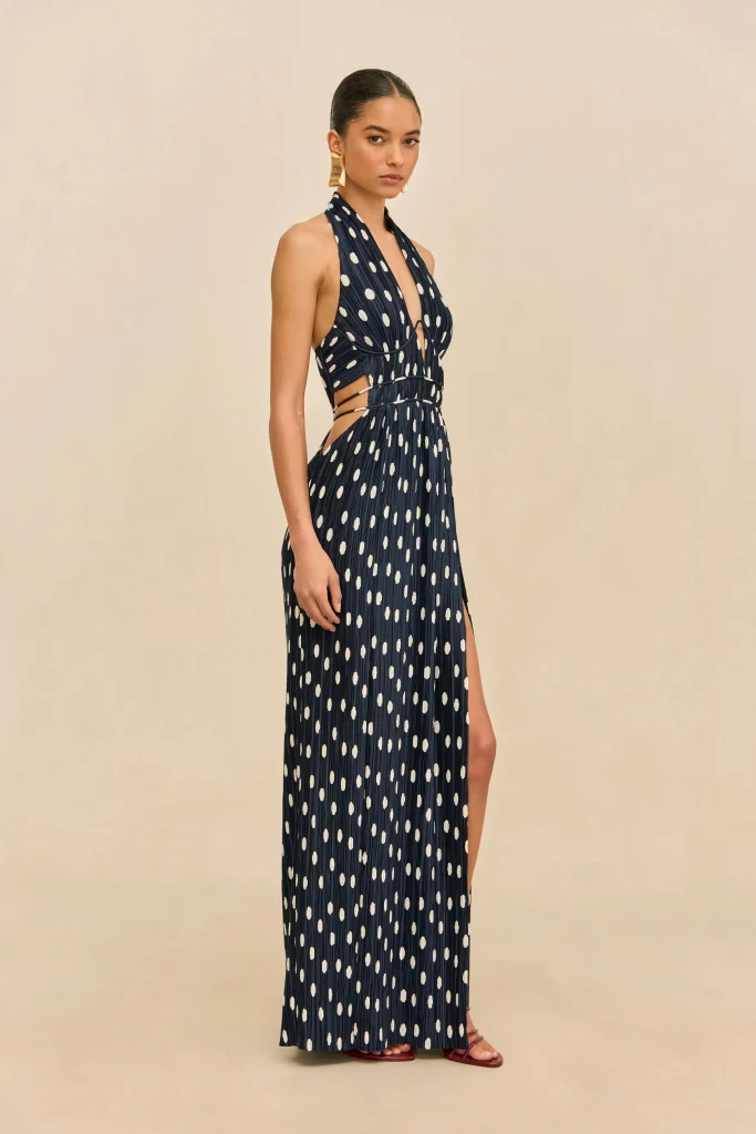 Model wearing a navy polka-dot halter maxi dress with side cutouts, standing against a beige backdrop.