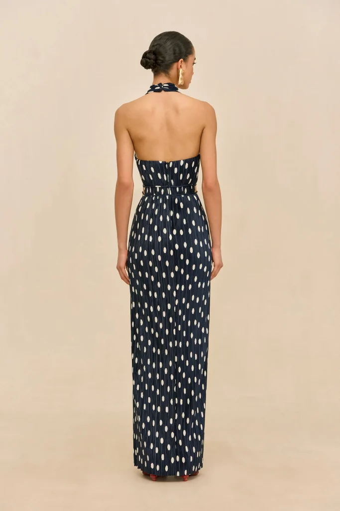 Back view of a woman in a navy polka-dot halter-neck maxi dress, hair in a bun, standing against a beige backdrop.