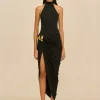 Female model wearing a black halter-neck gown with a draped skirt and gold waist accents, standing against a beige background.