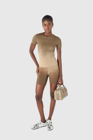 Woman in tan gradient matching athletic set (short-sleeve top and shorts) with a repeating pattern, holding a beige handbag and wearing white sneakers.