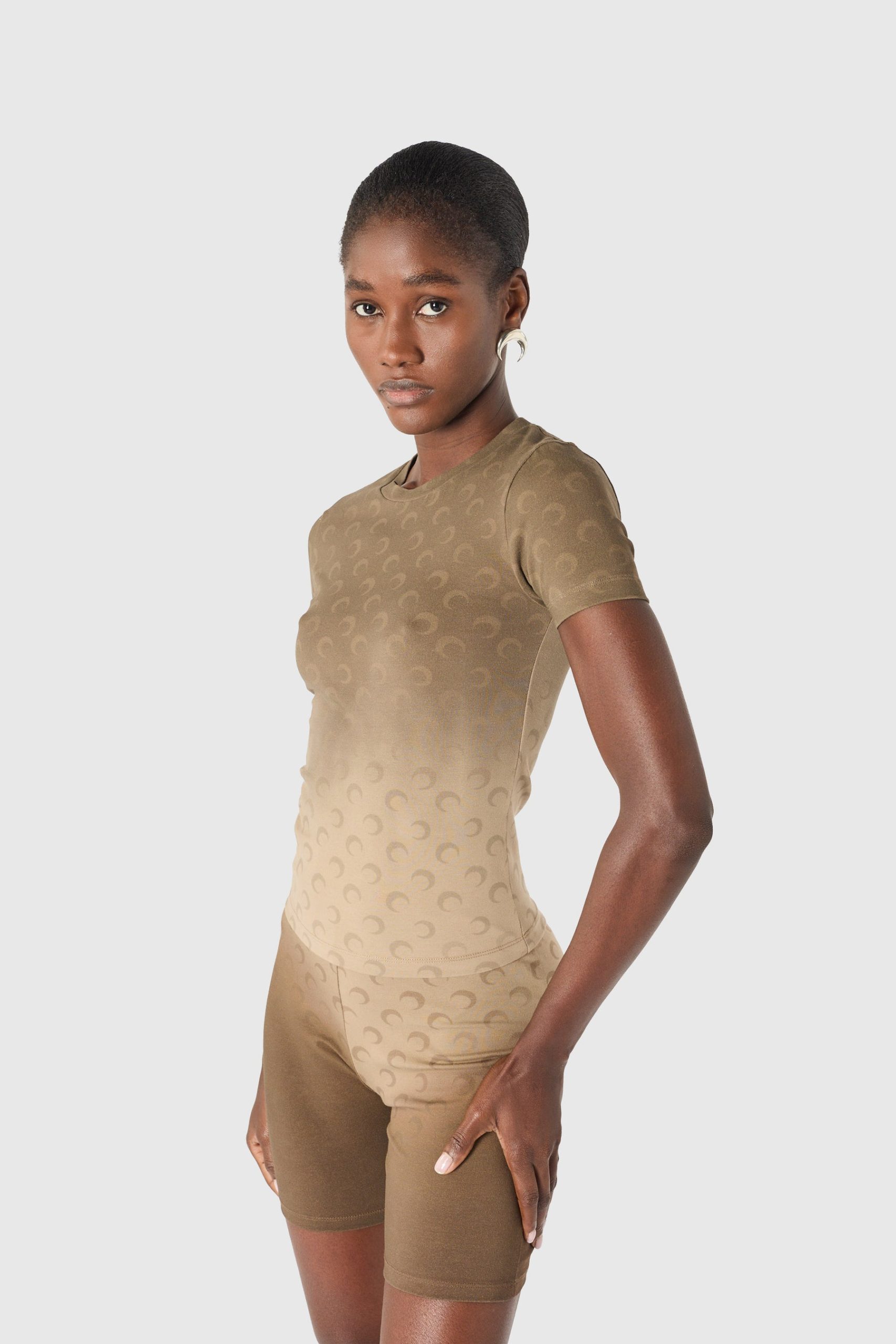 Woman modeling a beige seamless shapewear set (top and shorts) with a subtle pattern, standing in profile against a light gray backdrop.