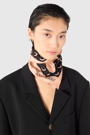 Close-up portrait of a woman in a black blazer, pale pink scarf with crescent pattern, and a gold hoop earring against a gray background.