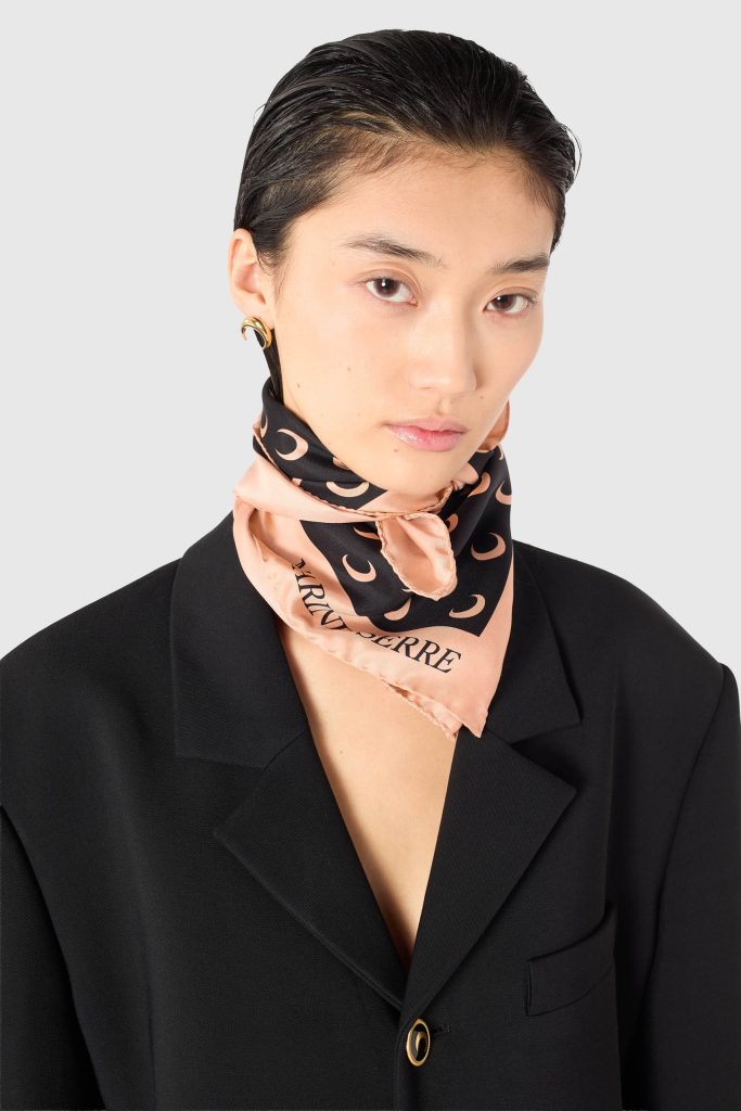 Close-up portrait of a woman in a black blazer, pale pink scarf with crescent pattern, and a gold hoop earring against a gray background.