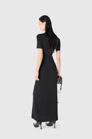 Woman in a long black dress standing with her back to the camera, holding a small handbag against a light gray background.
