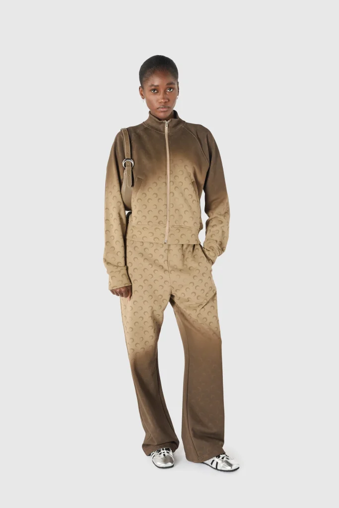 Model wearing a gradient brown monogram track jacket and matching pants, carrying a beige shoulder bag and wearing silver sneakers against a light gray background.