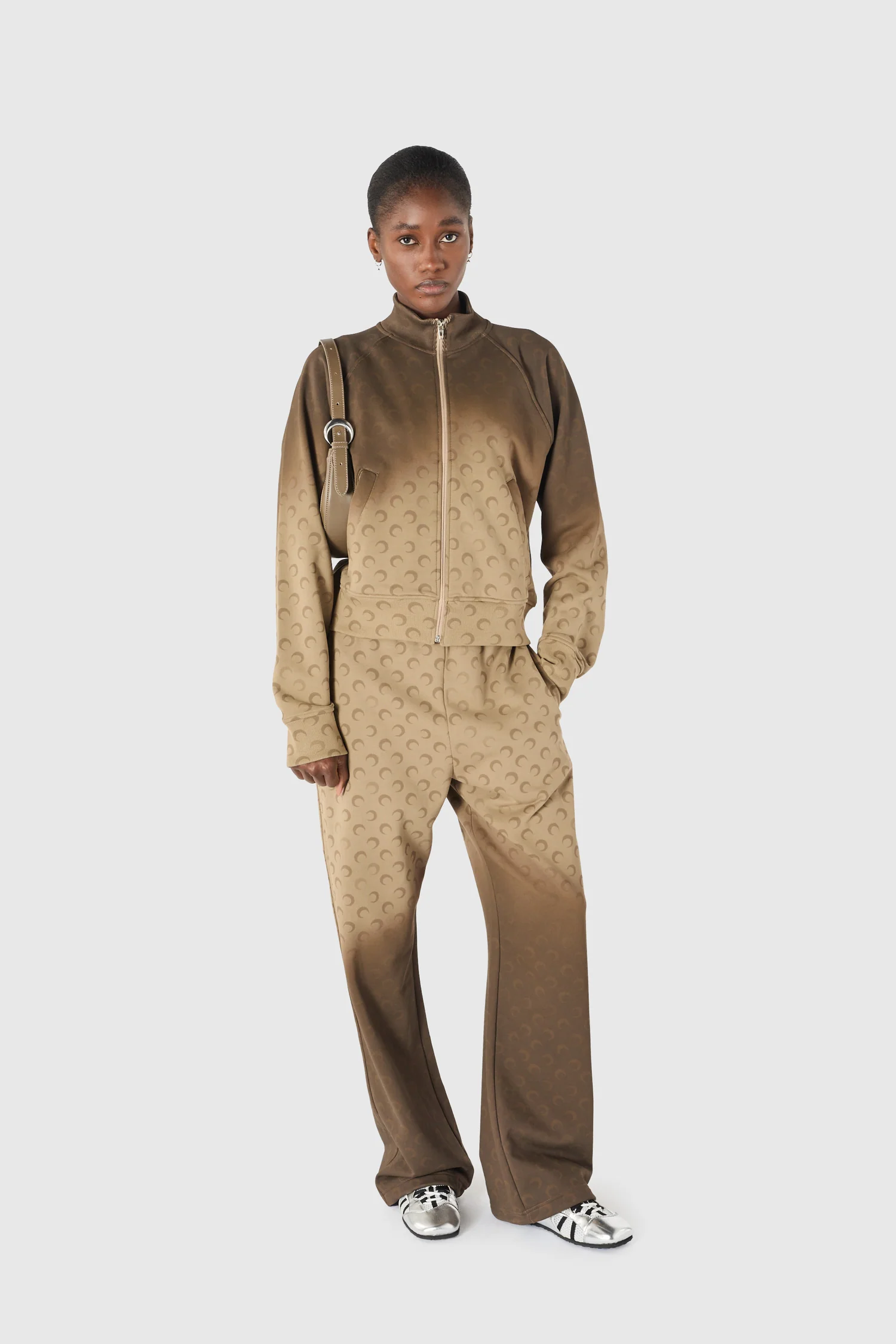 Model wearing a gradient brown monogram track jacket and matching pants, carrying a beige shoulder bag and wearing silver sneakers against a light gray background.