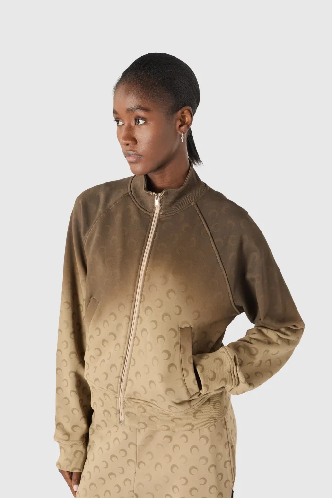 Portrait of a woman wearing a brown gradient zip-front jacket with a repeating pattern, against a light gray background—looking to the left.