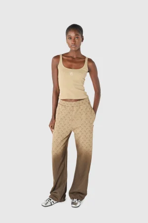 Full-body shot of a young woman wearing a beige tank top and tan patterned wide-leg pants, standing against a light gray background.