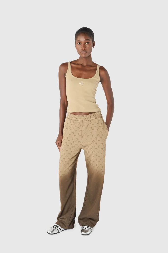 Full-body shot of a young woman wearing a beige tank top and tan patterned wide-leg pants, standing against a light gray background.