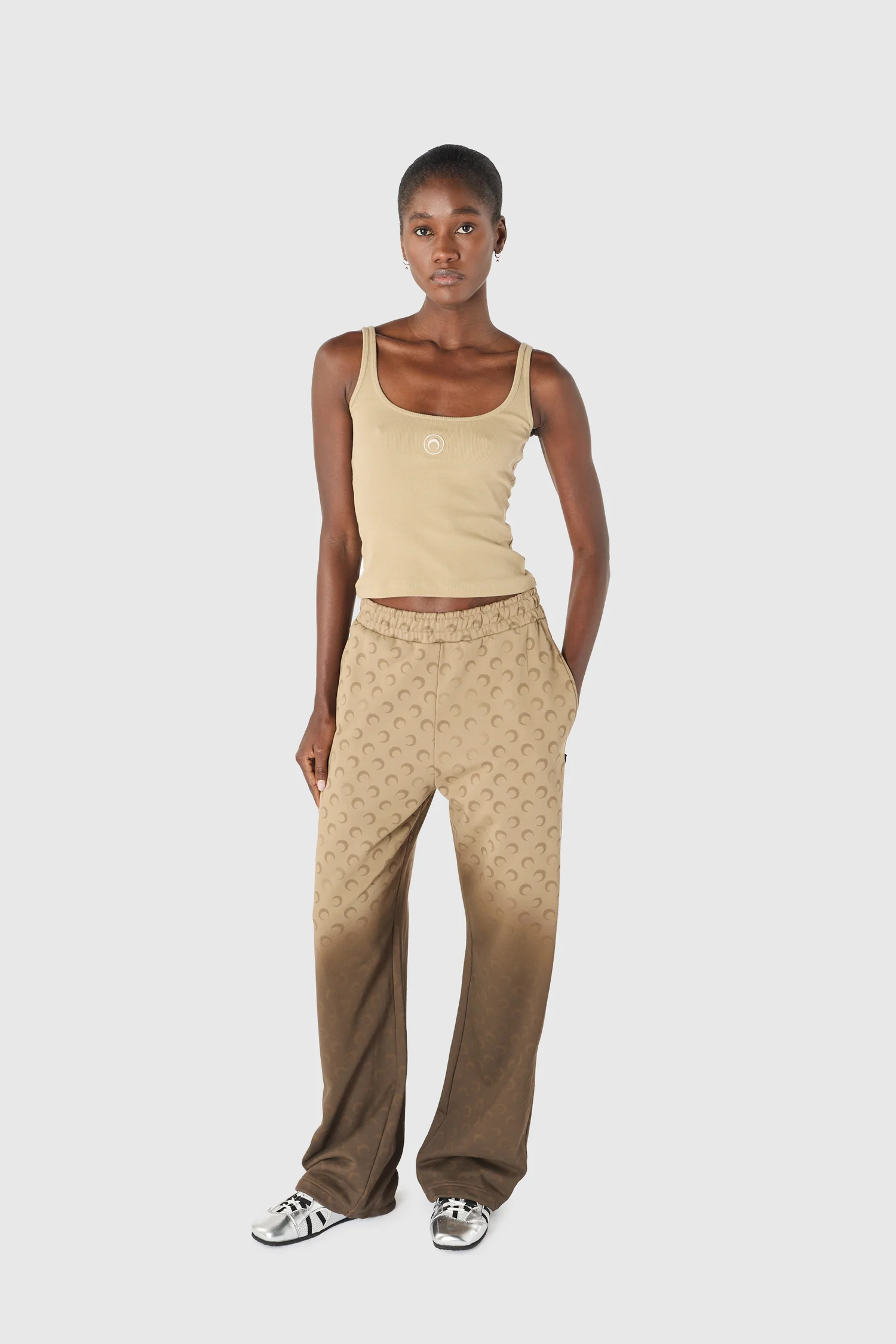 Full-body shot of a young woman wearing a beige tank top and tan patterned wide-leg pants, standing against a light gray background.