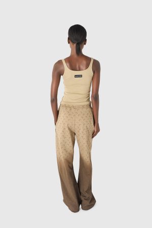 Back view of a person wearing a beige tank top and tan patterned pants, with a small label on the tank top's back and hair tied in a low ponytail.