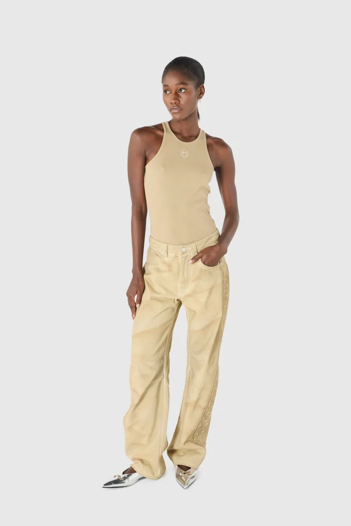 Full-body shot of a woman wearing a beige tank top and matching beige cargo pants, standing with one hand in pocket against a light gray background.