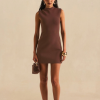 Woman wearing a sleeveless brown mini dress, holding a small textured brown handbag, with gold earrings and bracelets, standing against a beige backdrop.