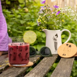 Glass of pink juice with a lime wheel on a cork coaster, with sunglasses, a white floral pitcher, and coasters on a weathered wooden table in a garden setting.