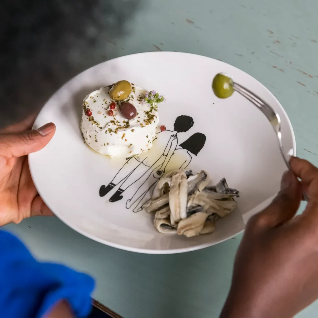 A plate holds a small round herb-crusted dessert topped with olives and flowers, with mushroom pasta on the side and a fork picking up a green olive.
